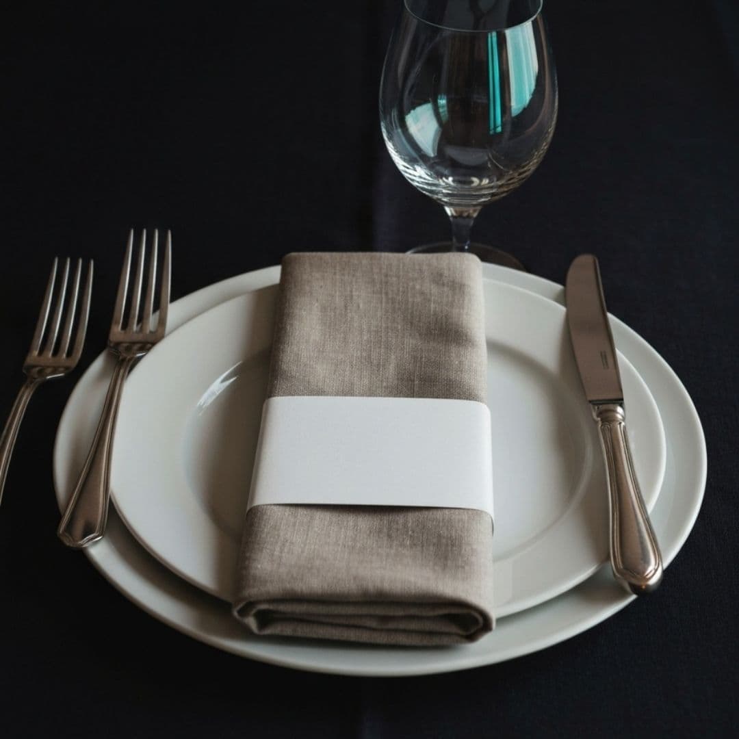 A single place setting on dark linen: a white porcelain plate with a folded napkin in a plain paper band, silver forks and knife either side, and an empty crystal wine glass behind.