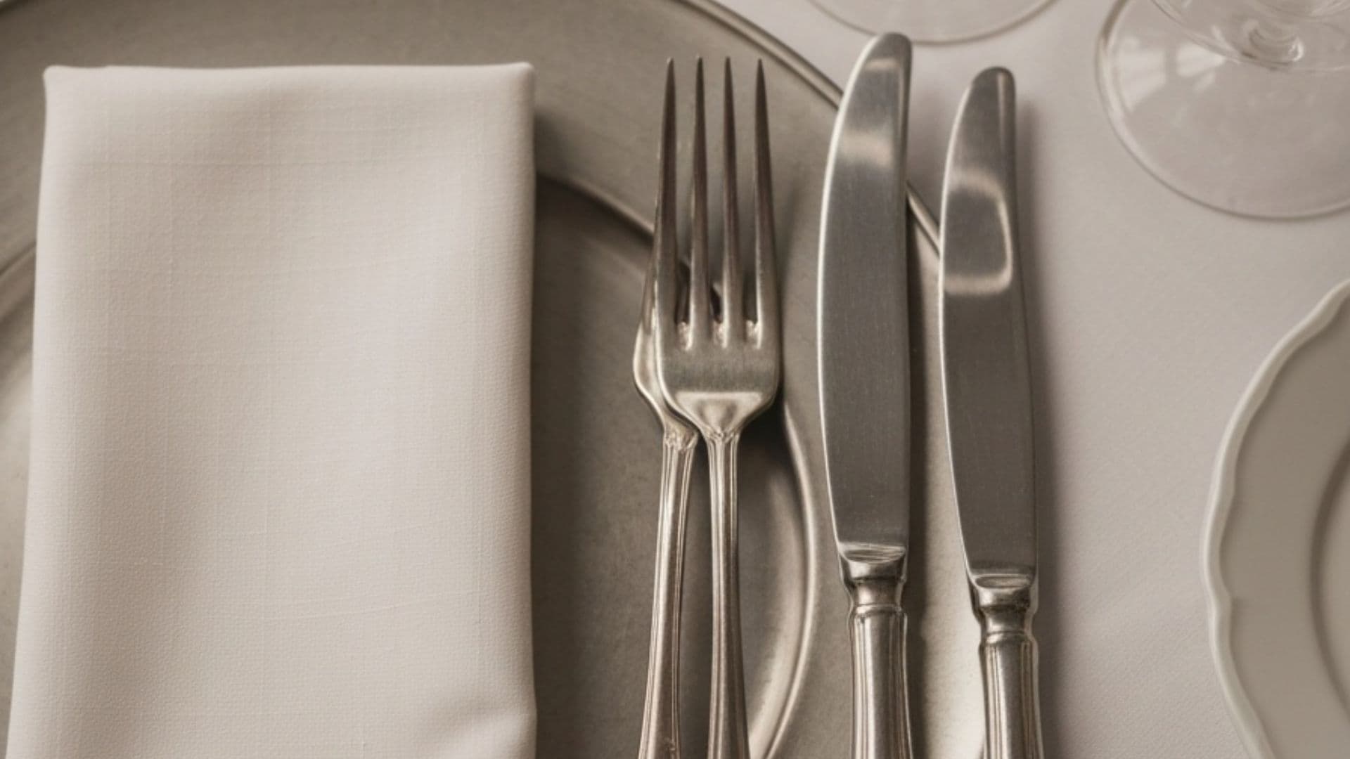 An overhead close-up of a formal place setting: folded linen napkin, silver fork and knives, charger plate and a wine glass on linen.