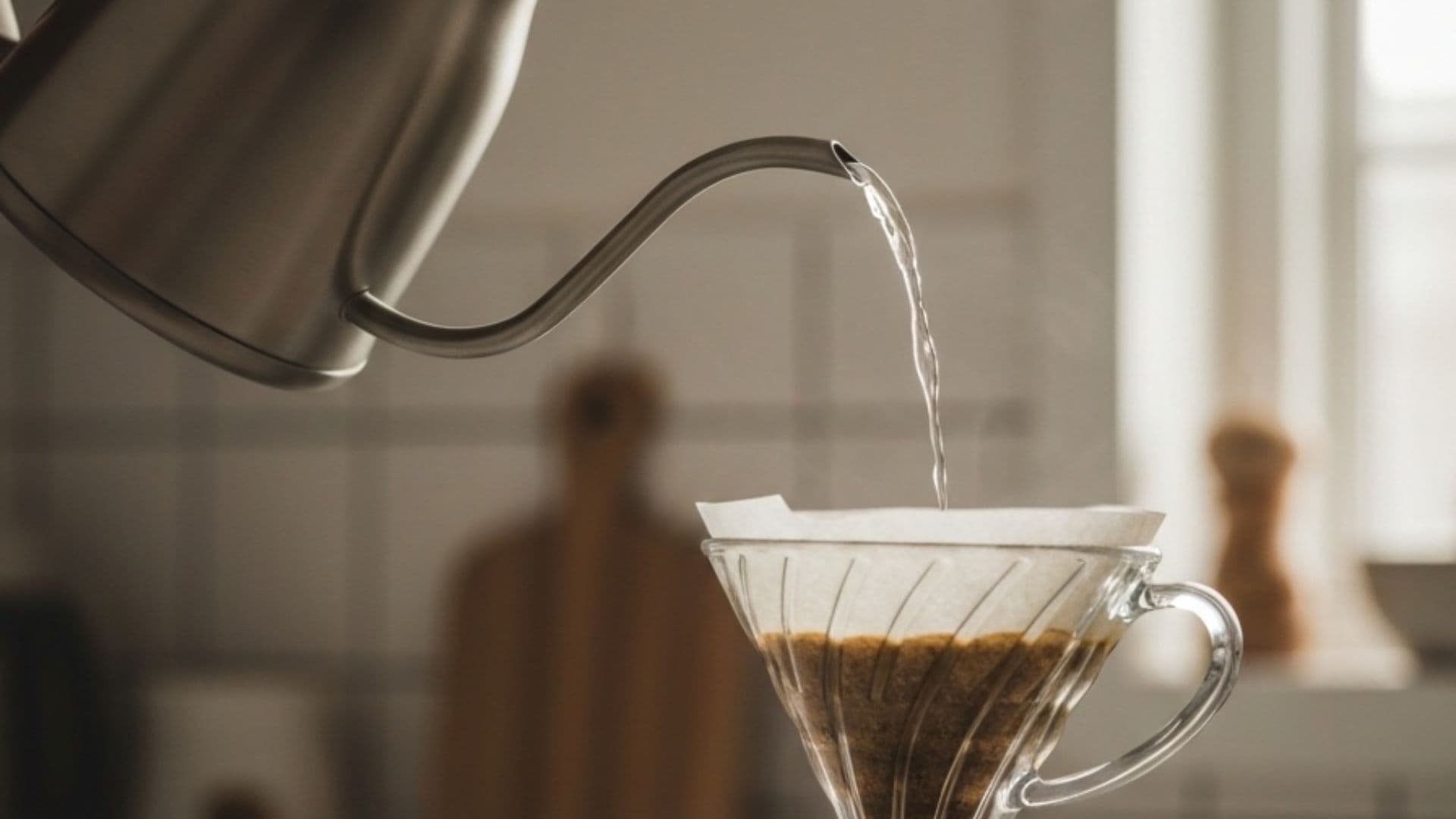 A gooseneck pour-over kettle mid-pour, a thin stream of water arcing from its narrow spout into a glass dripper in warm window light.