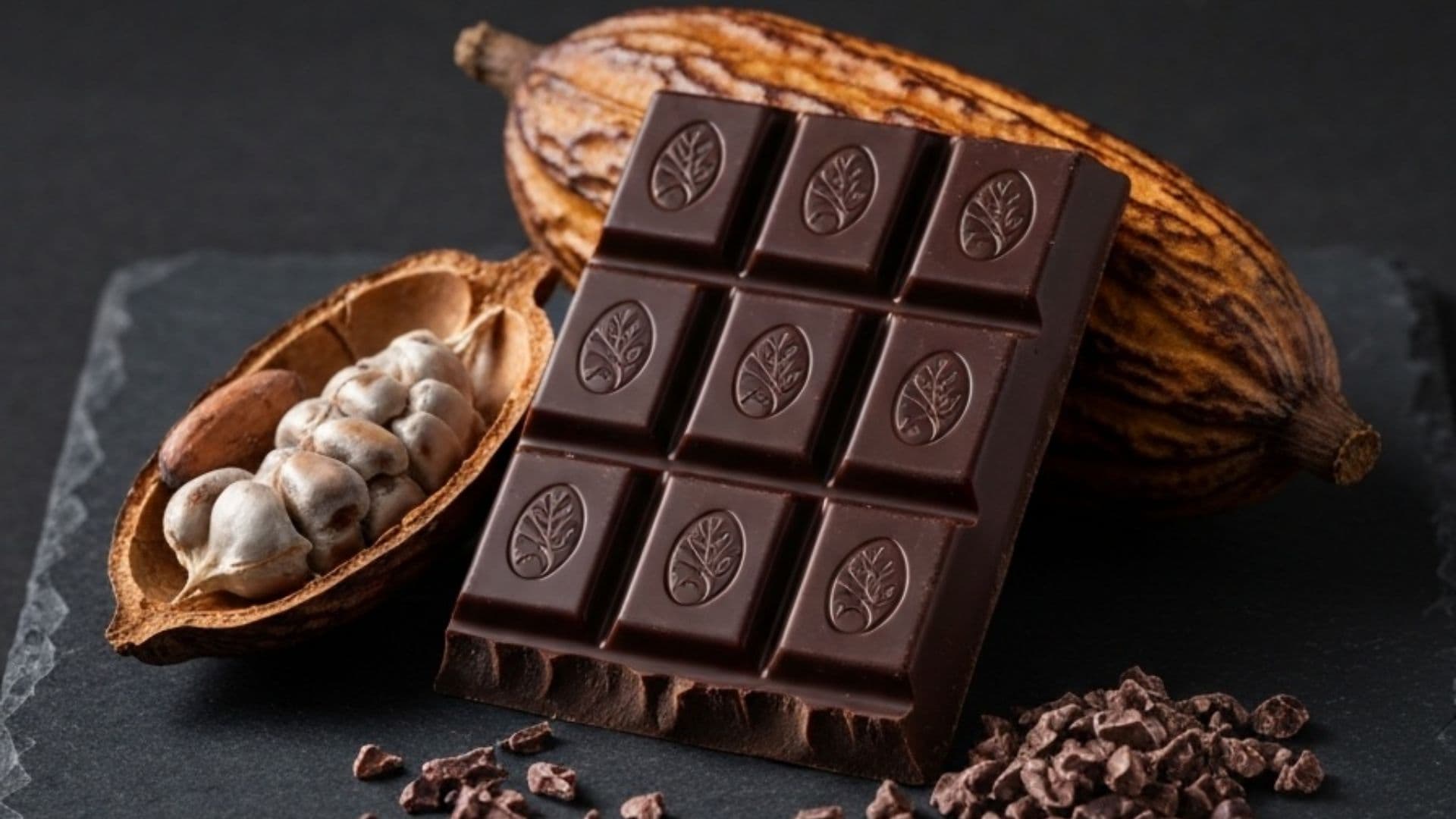 A dark single-origin chocolate bar beside a split cacao pod with beans and scattered nibs on slate.